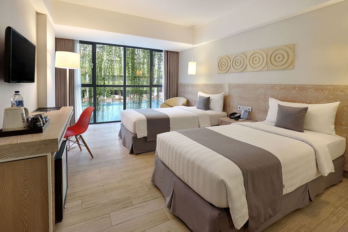 A hotel room with two beds, a desk with a chair, a TV, and a large window with a view of greenery.