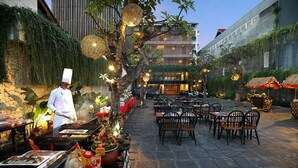 Uma área de refeições ao ar livre com mesas e cadeiras, um chef preparando comida e um edifício com design moderno ao fundo.