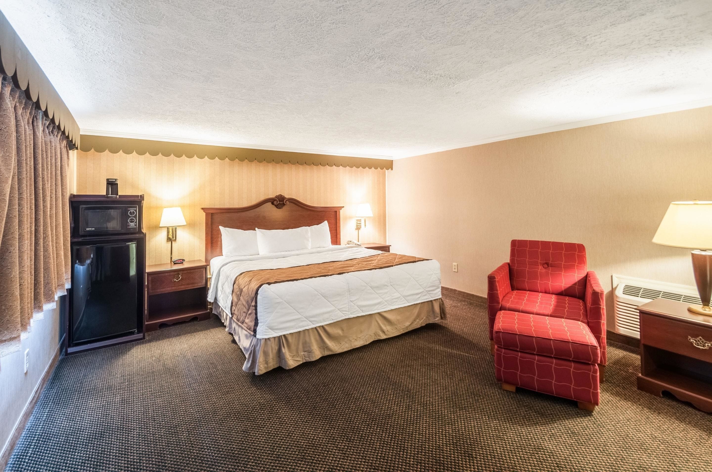 A hotel room with a bed, a red armchair, a desk, a lamp, a microwave, and a nightstand.
