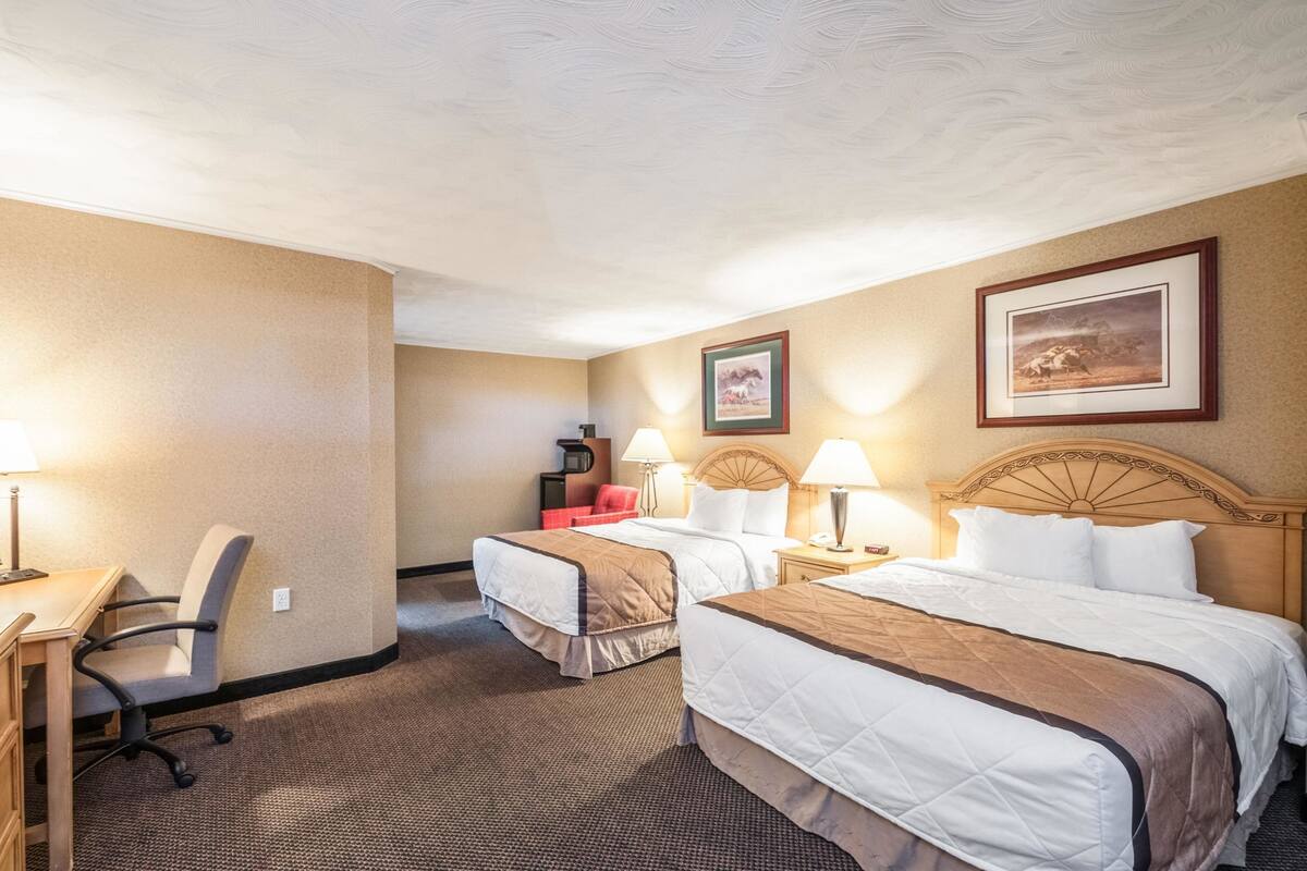 A hotel room with two beds, a desk, and a chair. There are lamps on the nightstands and a framed picture on the wall.