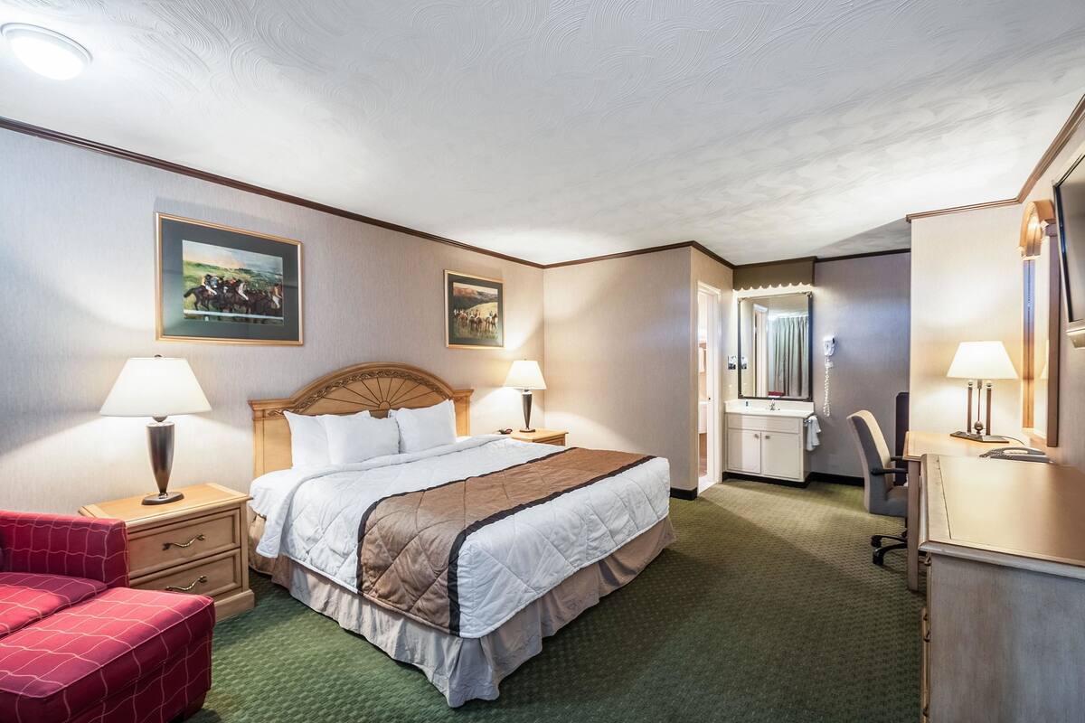 A hotel room with a large bed, two bedside lamps, a desk with a chair, a mirror, and a red armchair.