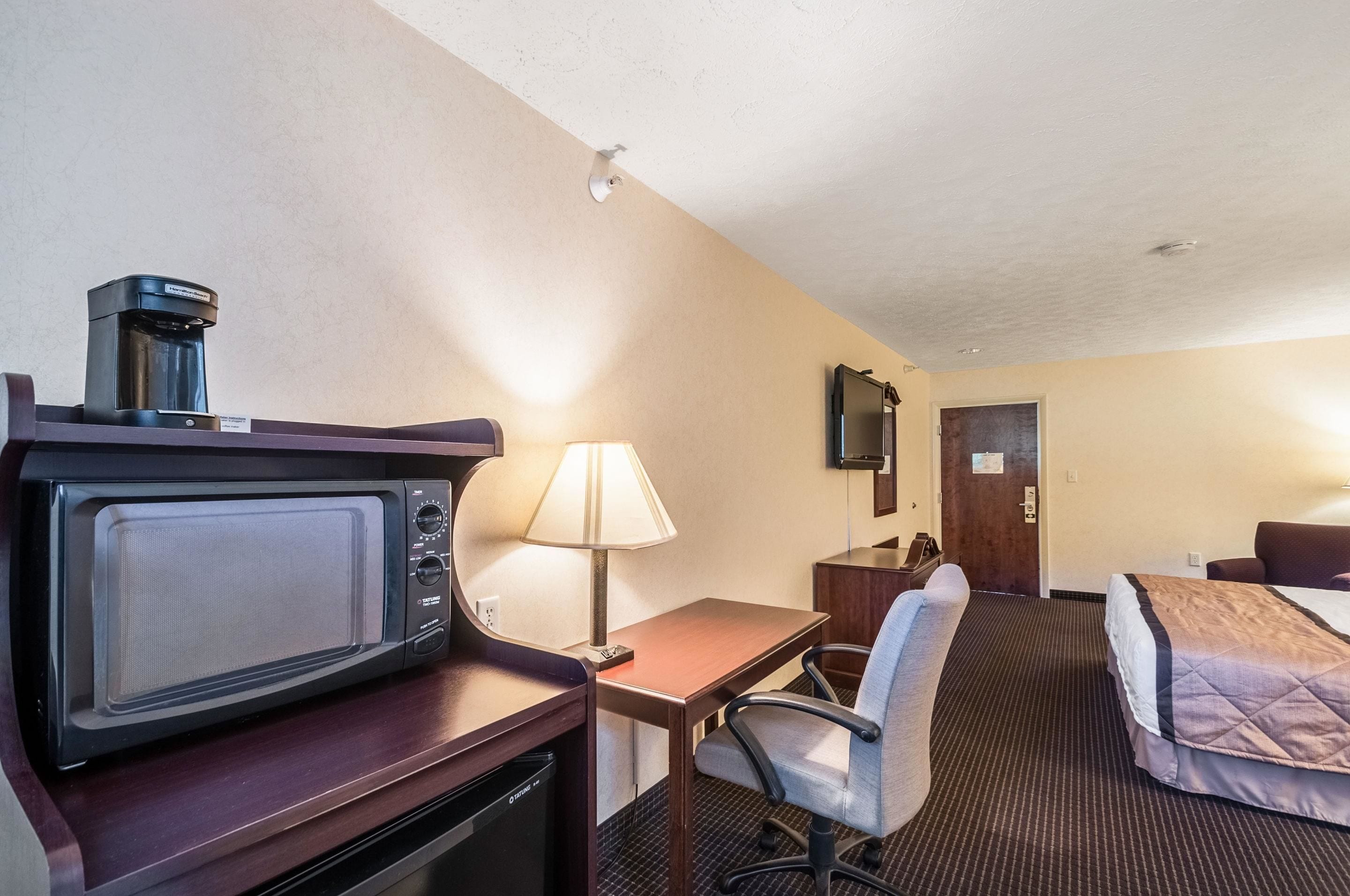 A hotel room with a microwave, a lamp, a desk, a chair, a bed, and a TV mounted on the wall.