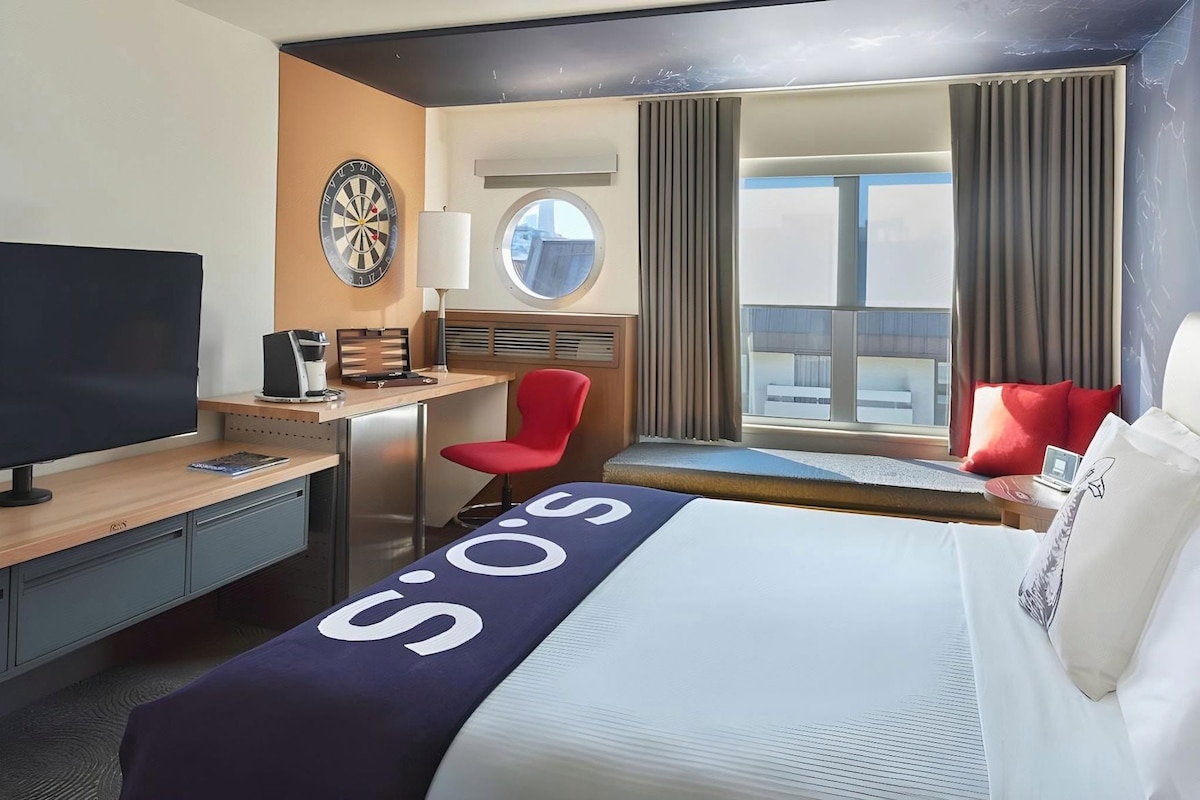 Une chambre d’hôtel avec un lit, un bureau, une télévision, un jeu de fléchettes et une fenêtre avec des rideaux.