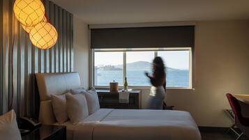 A hotel room with a bed, a nightstand, a lamp, a chair, and a window with a view of the sea.
