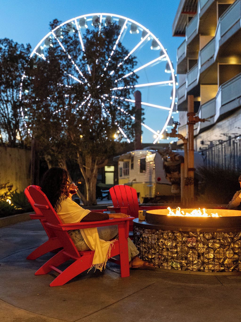 An outdoor seating area with red chairs and a fire pit, a Ferris wheel in the background, and a building with balconies.