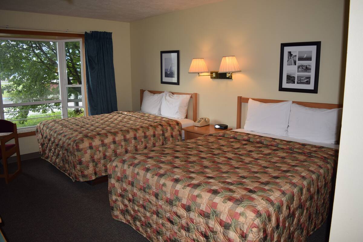 A hotel room with two beds, a desk, and a chair. There are two wall-mounted lamps and framed pictures on the wall.
