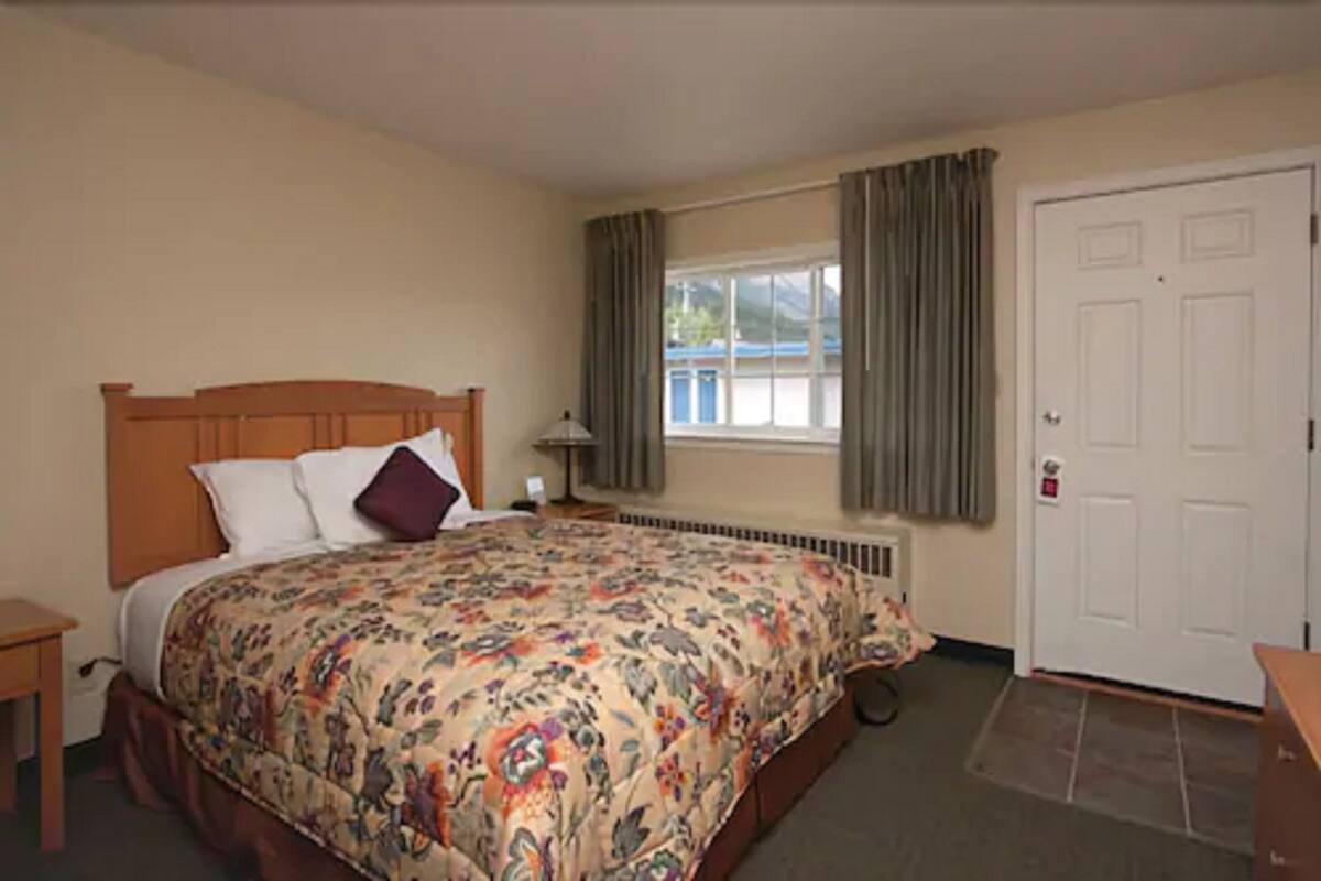 A bedroom with a large bed, a nightstand, a window with curtains, a radiator, and a door.