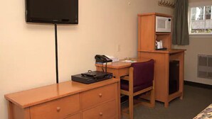 Habitación de hotel con una televisión montada en la pared, un escritorio de madera con un teléfono y un pequeño dispositivo electrónico, una silla y un microondas en un mueble de madera.