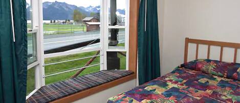 A bedroom with a bed, a window with a view of a road and mountains, and a bench outside the window.