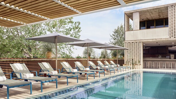 Area piscina con lettini, ombrelloni, un bar e un edificio con un balcone.
