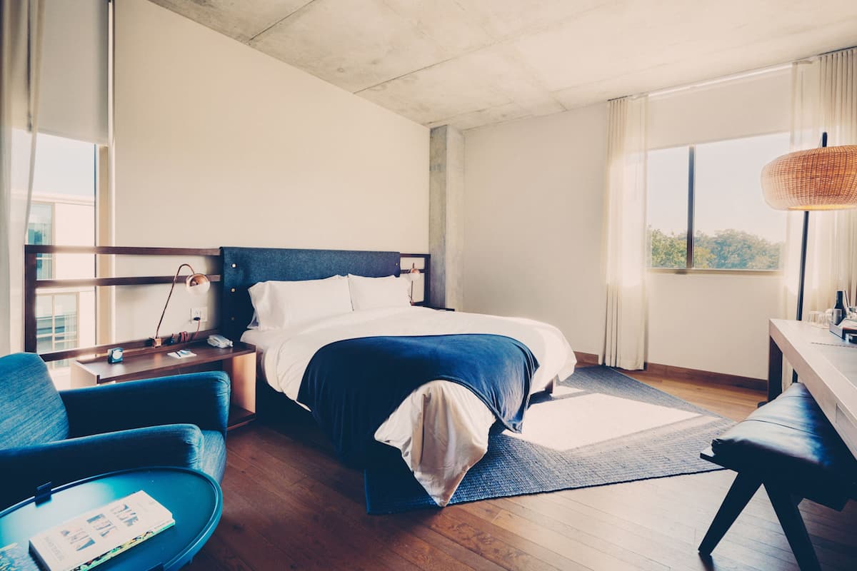 Quarto de hotel com cama, escrivaninha, cadeira e janela com cortinas.