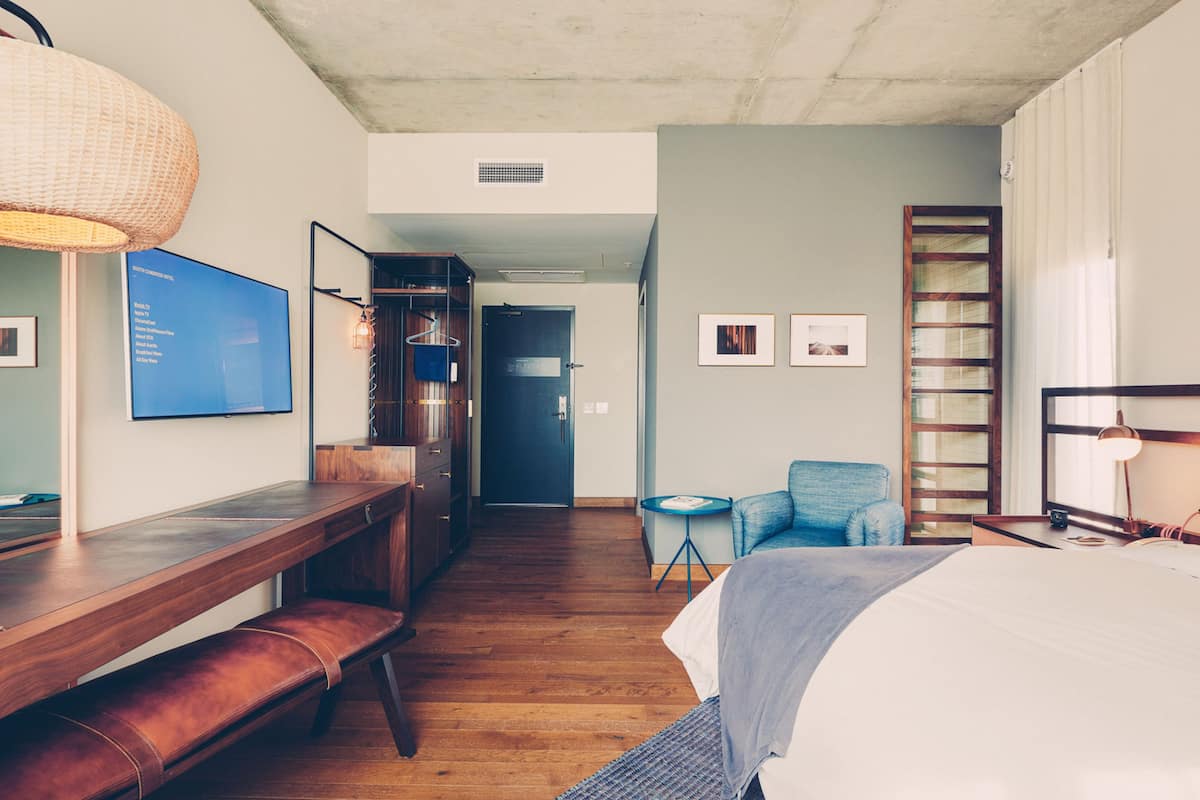 Quarto de hotel com secretária de madeira, uma cama, poltrona azul e mesa redonda pequena.