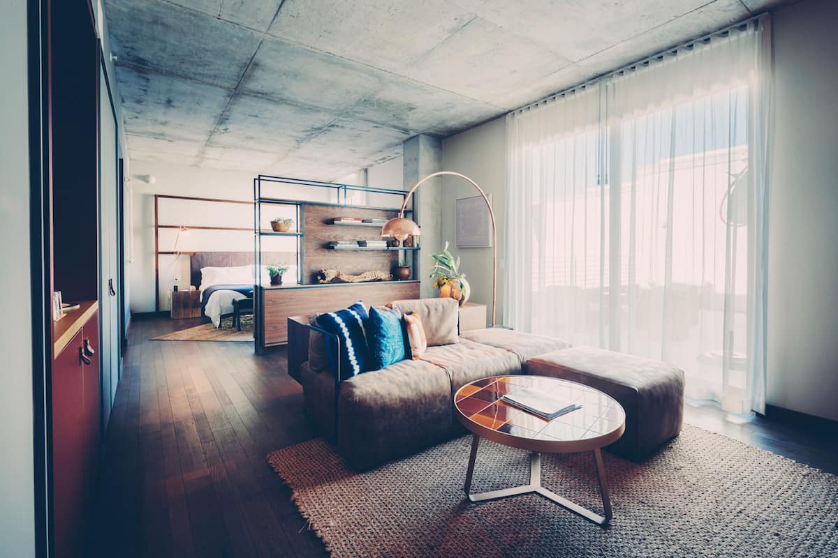 Uma sala de estar moderna com sofá, apoio para os pés e mesa de centro. Há uma estante com livros e objetos decorativos, além de uma luminária. O ambiente possui janelas amplas com cortinas leves.