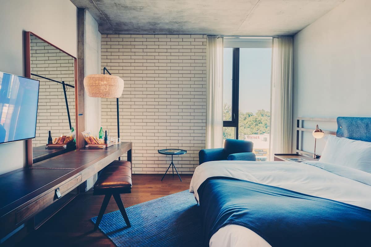 Quarto de hotel moderno com uma cama grande, uma escrivaninha com televisão de tela plana, uma mesinha redonda e uma janela ampla com vista para o exterior.