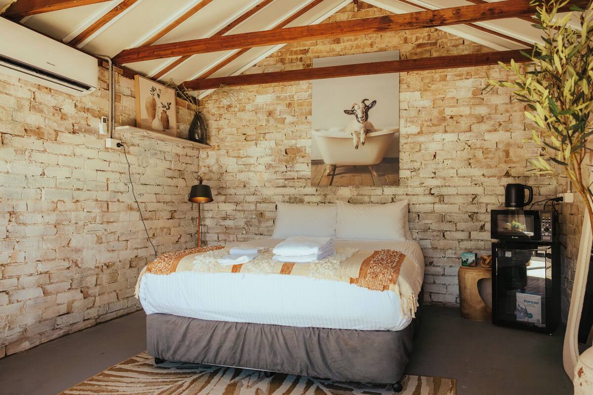 A room with a brick wall, a bed with white and brown bedding, a mounted picture of a goat in a bathtub, a small wooden side table, and a black coffee machine.