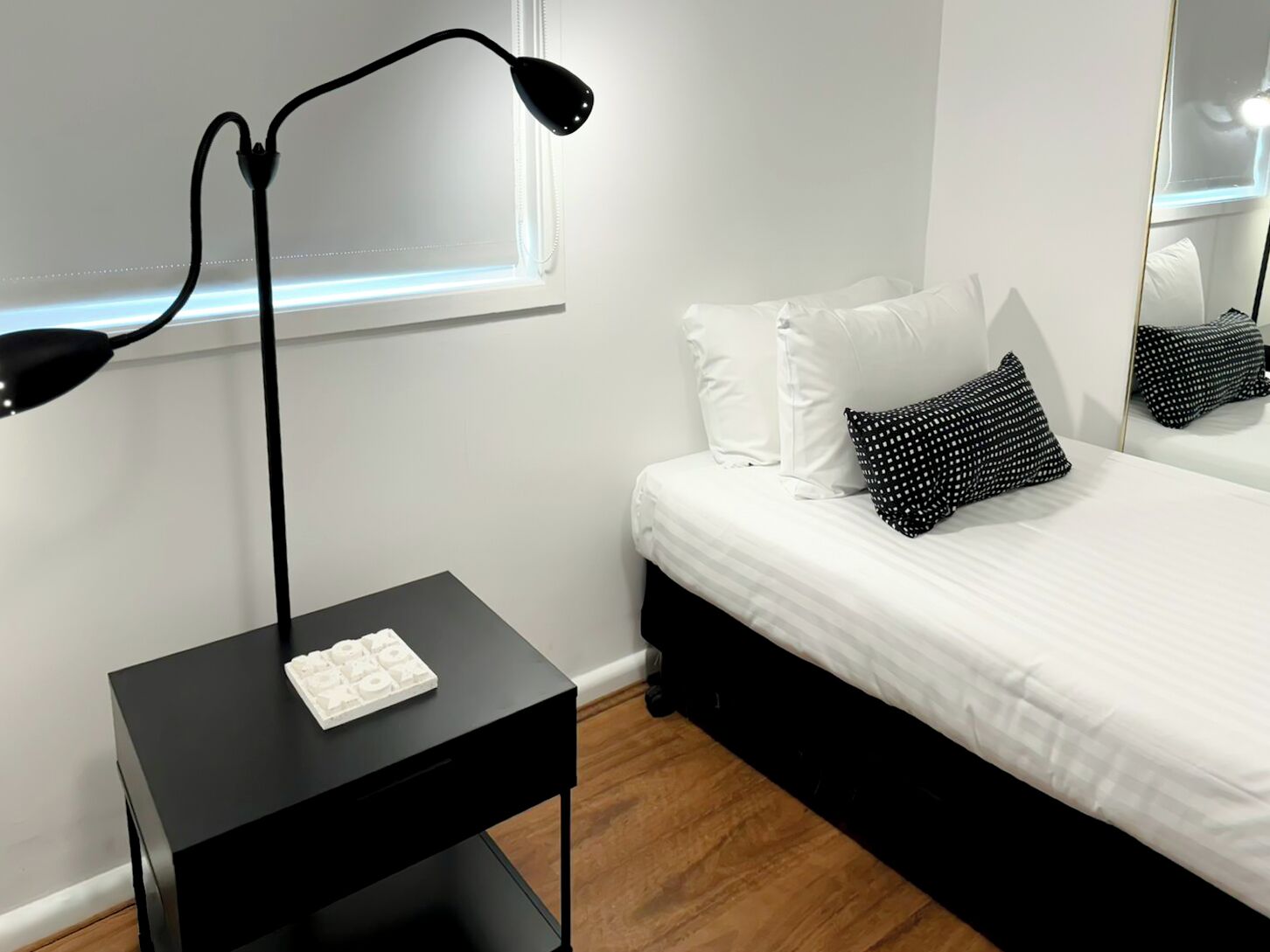 A bedroom with a black nightstand, a white bed with black and white patterned pillows, and a large mirror on the wall.