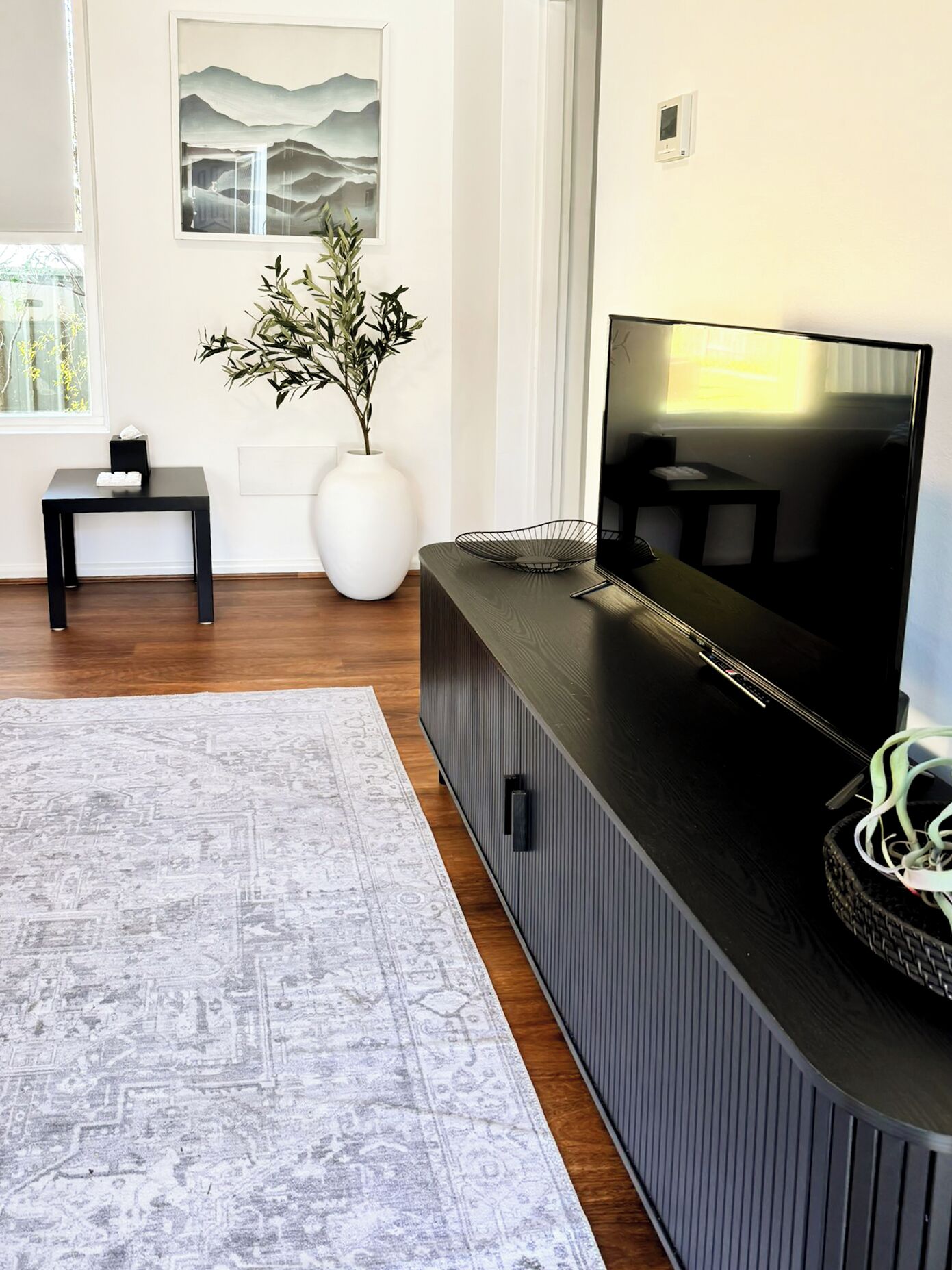 A modern living room with a large flat-screen TV, a white vase with greenery, and a patterned rug.