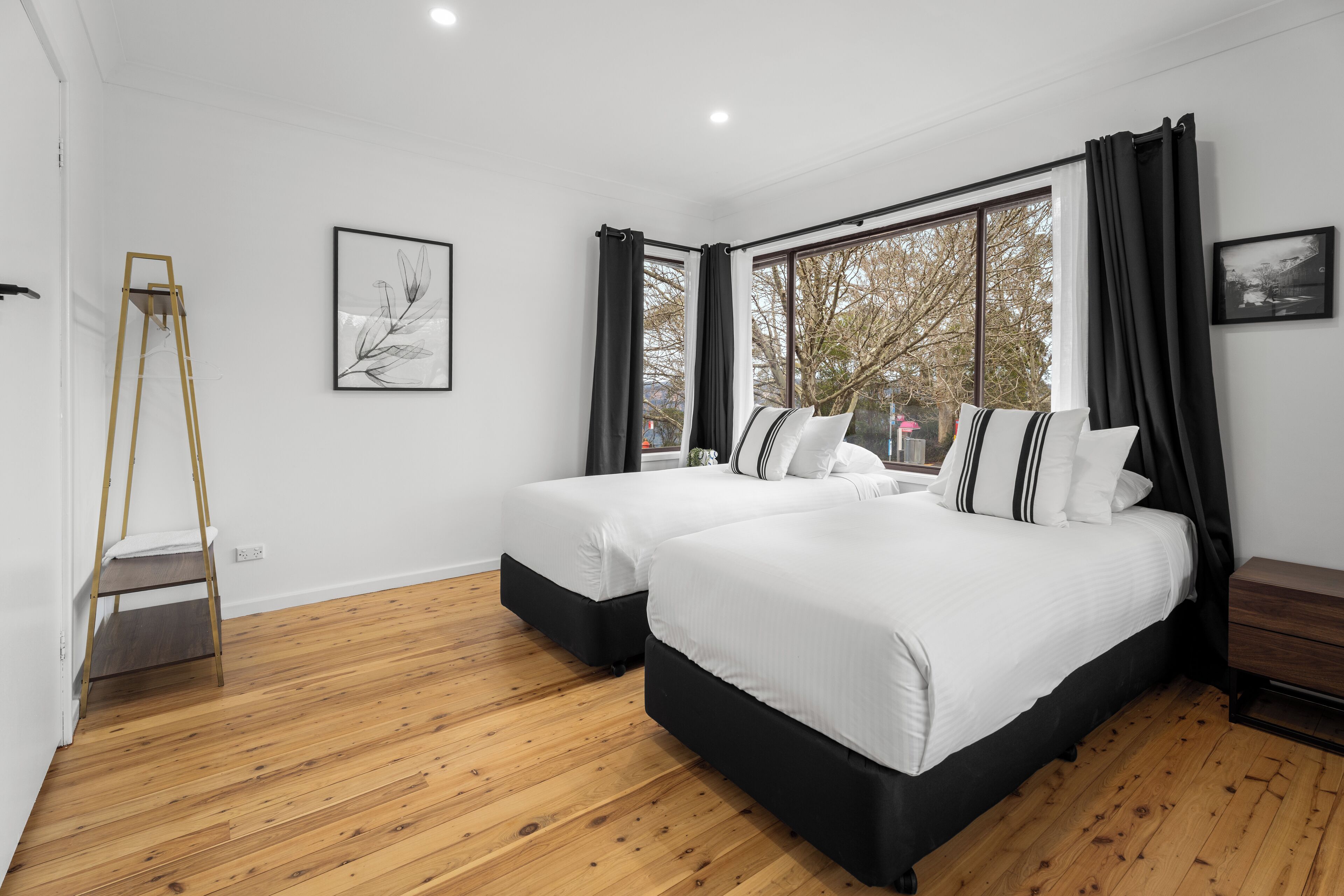 A modern bedroom with two beds, wooden flooring, and large windows with black curtains.