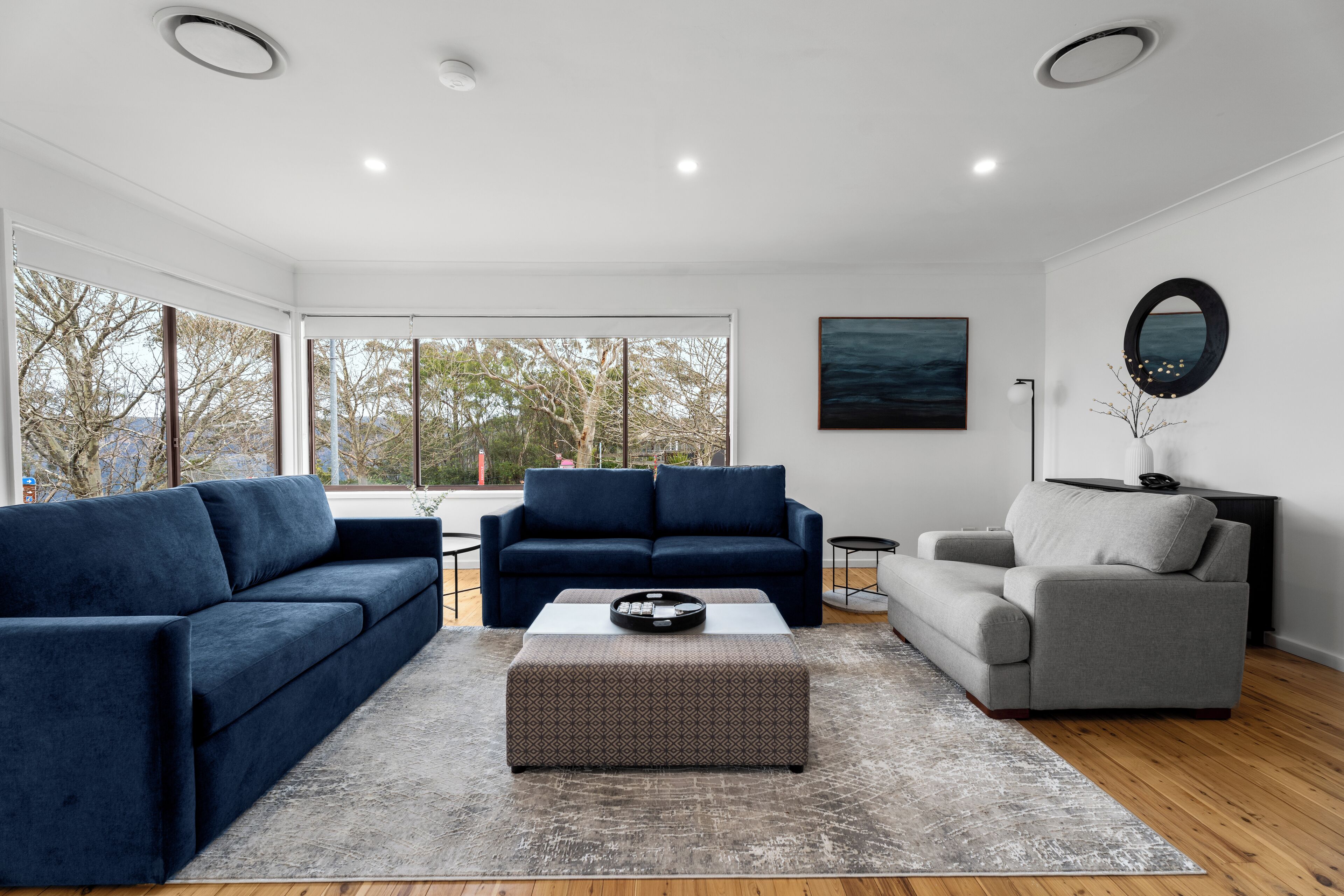 Een moderne woonkamer met een blauwe bank, een grijze bank en een gestreept voetenbankje. Er is een groot raam met uitzicht op bomen en een gebouw.