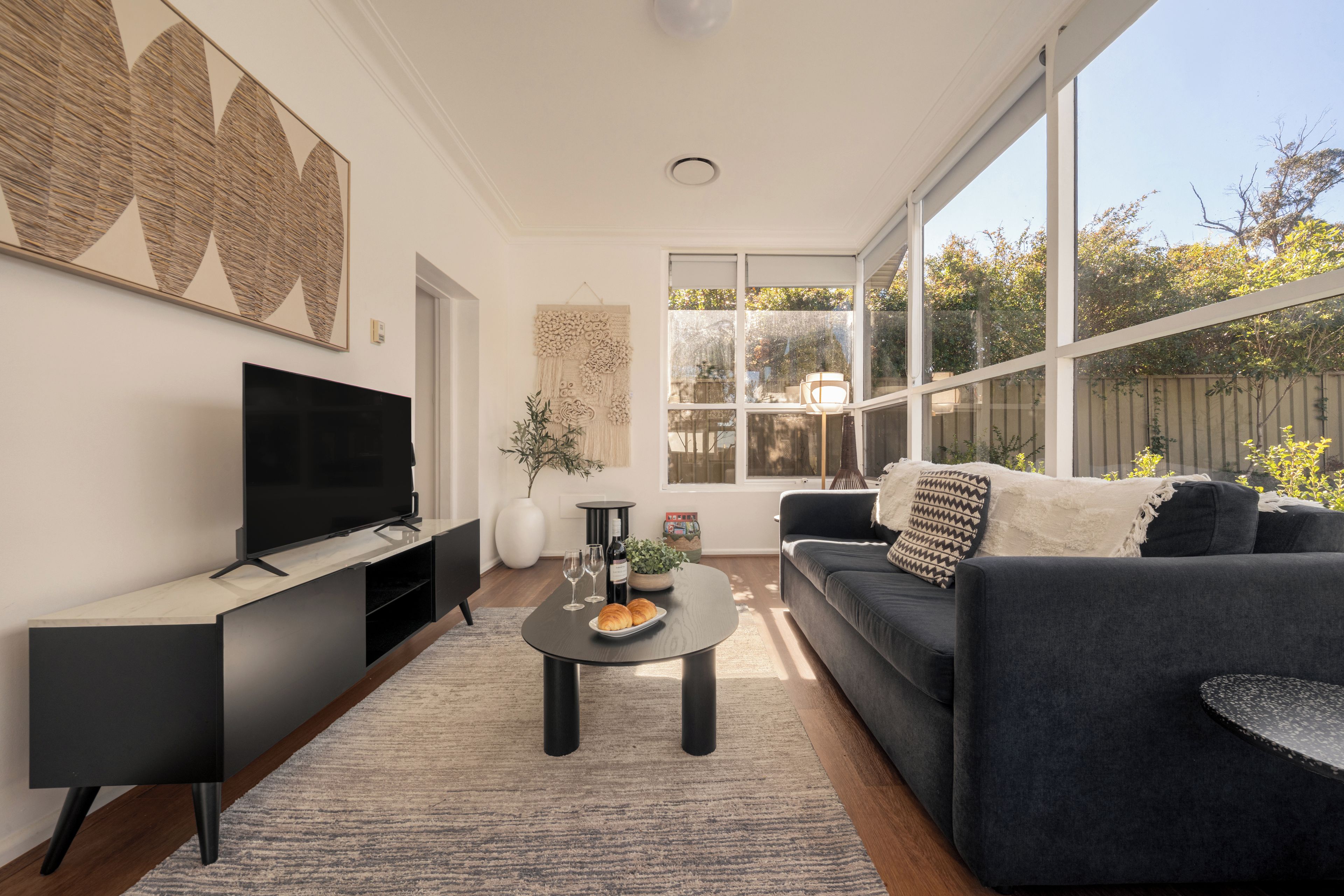 Ein modernes Wohnzimmer mit Fernseher, einer Couch, einem Couchtisch und einem großen Fenster mit Blick ins Freie.