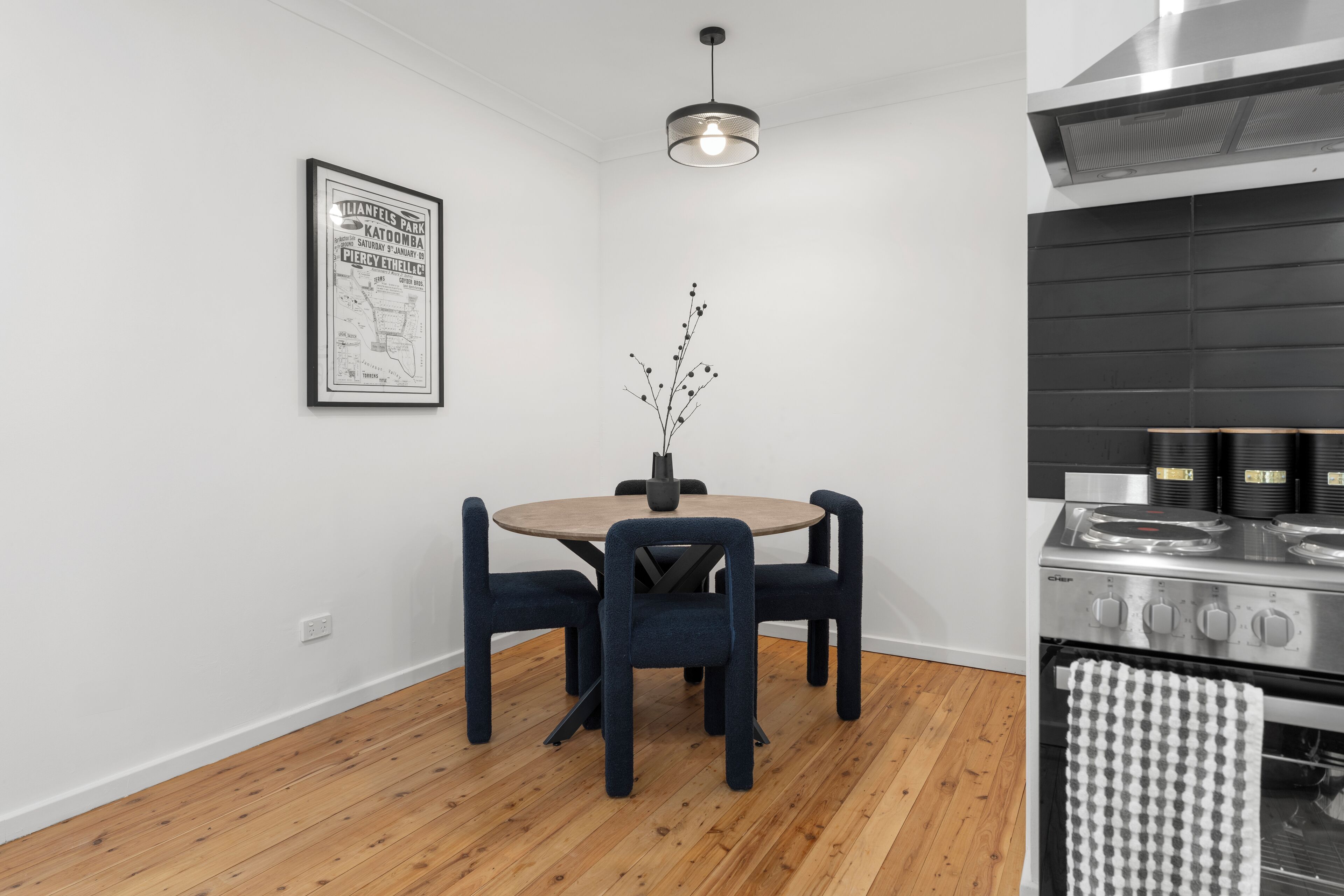 A modern kitchen with a round wooden table, dark blue chairs, a hanging light fixture, and a stainless steel stove.