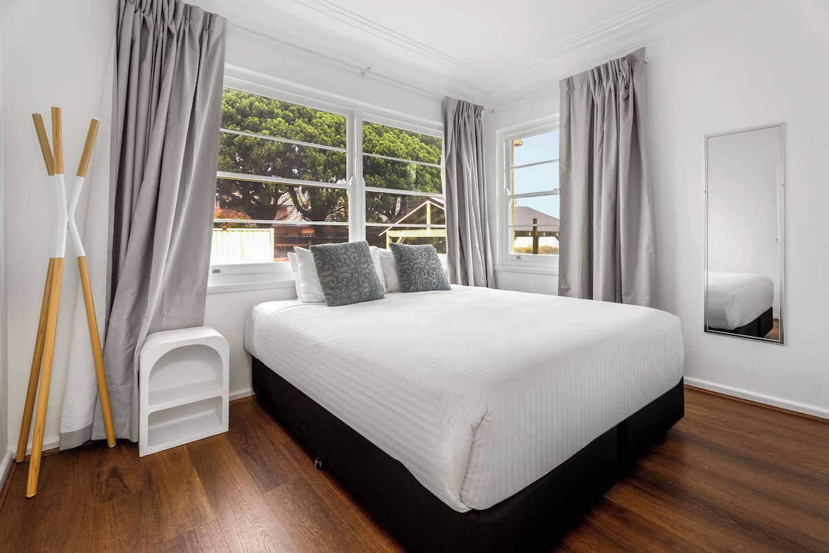 A bedroom with a large bed, a small white stool, a wooden ladder, and a window with curtains.