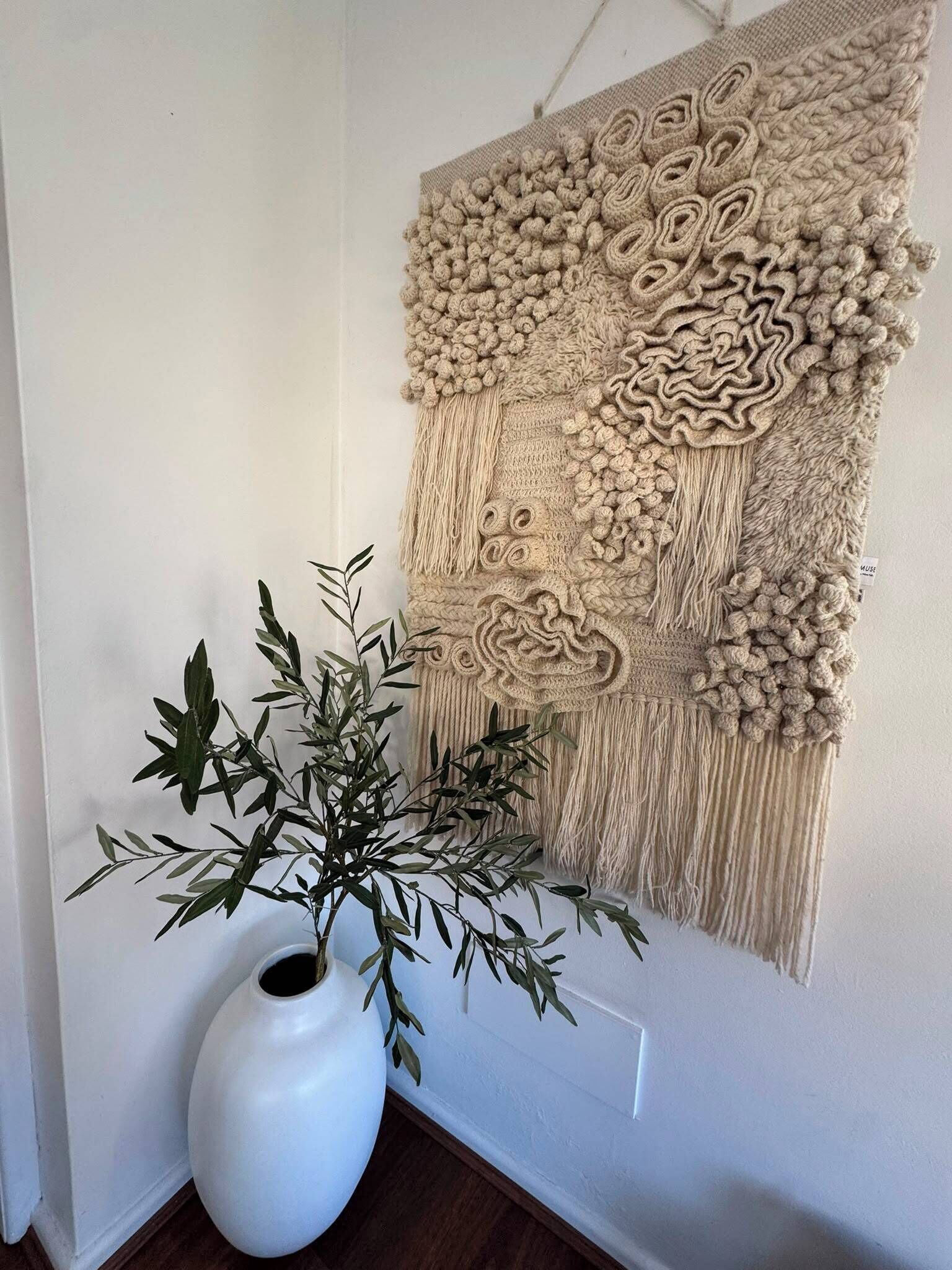 A wall-mounted macrame piece with a white vase and green foliage beside it.