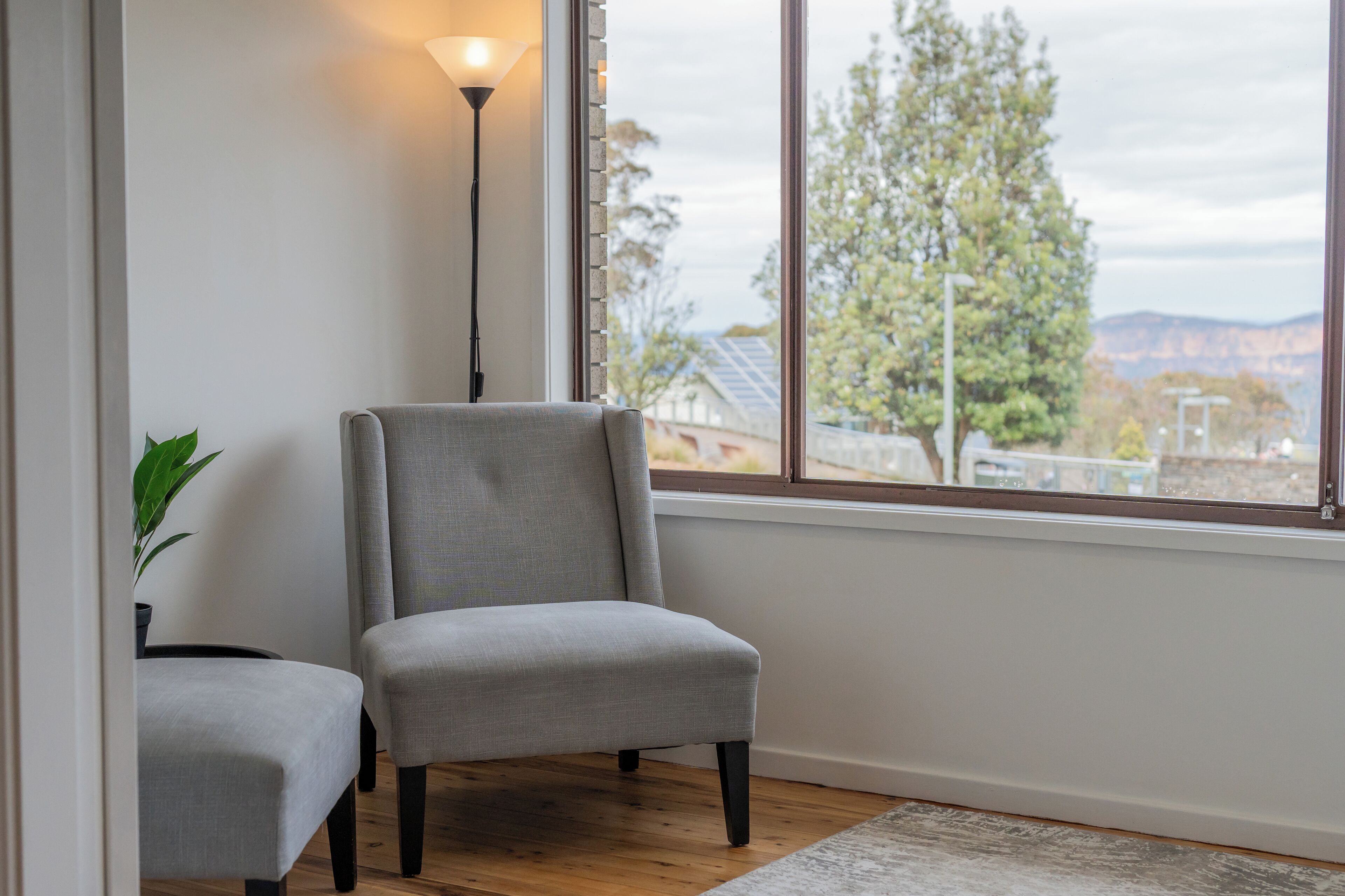 Una habitación con un sillón gris, una lámpara de pie y vistas a árboles y edificios a través de un ventanal grande.