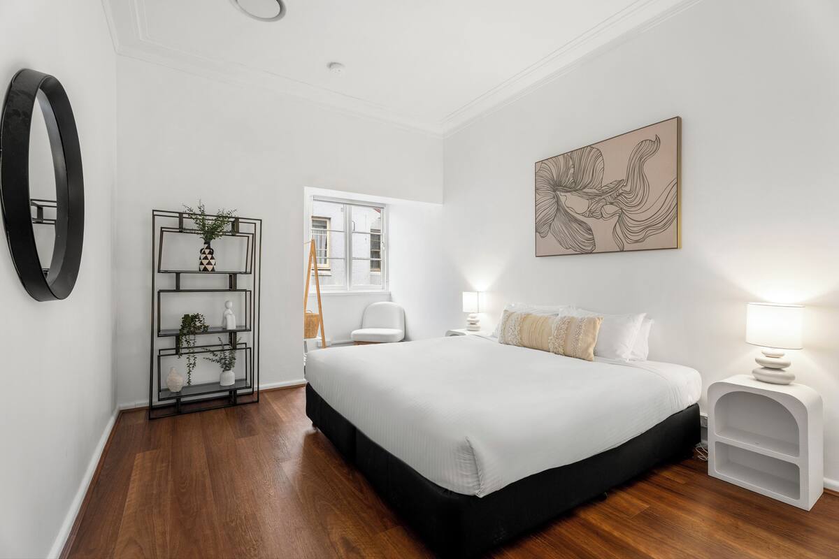 A bedroom with a large bed, a bedside table with a lamp, a wall-mounted mirror, and a decorative shelf with plants.