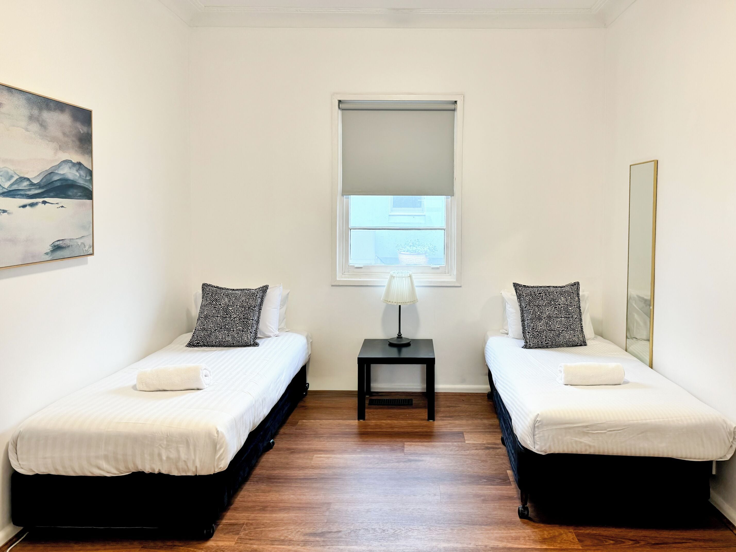 Una habitación pequeña y limpia con dos camas, una ventana con cortina, una mesita de noche con lámpara y un cuadro en la pared.