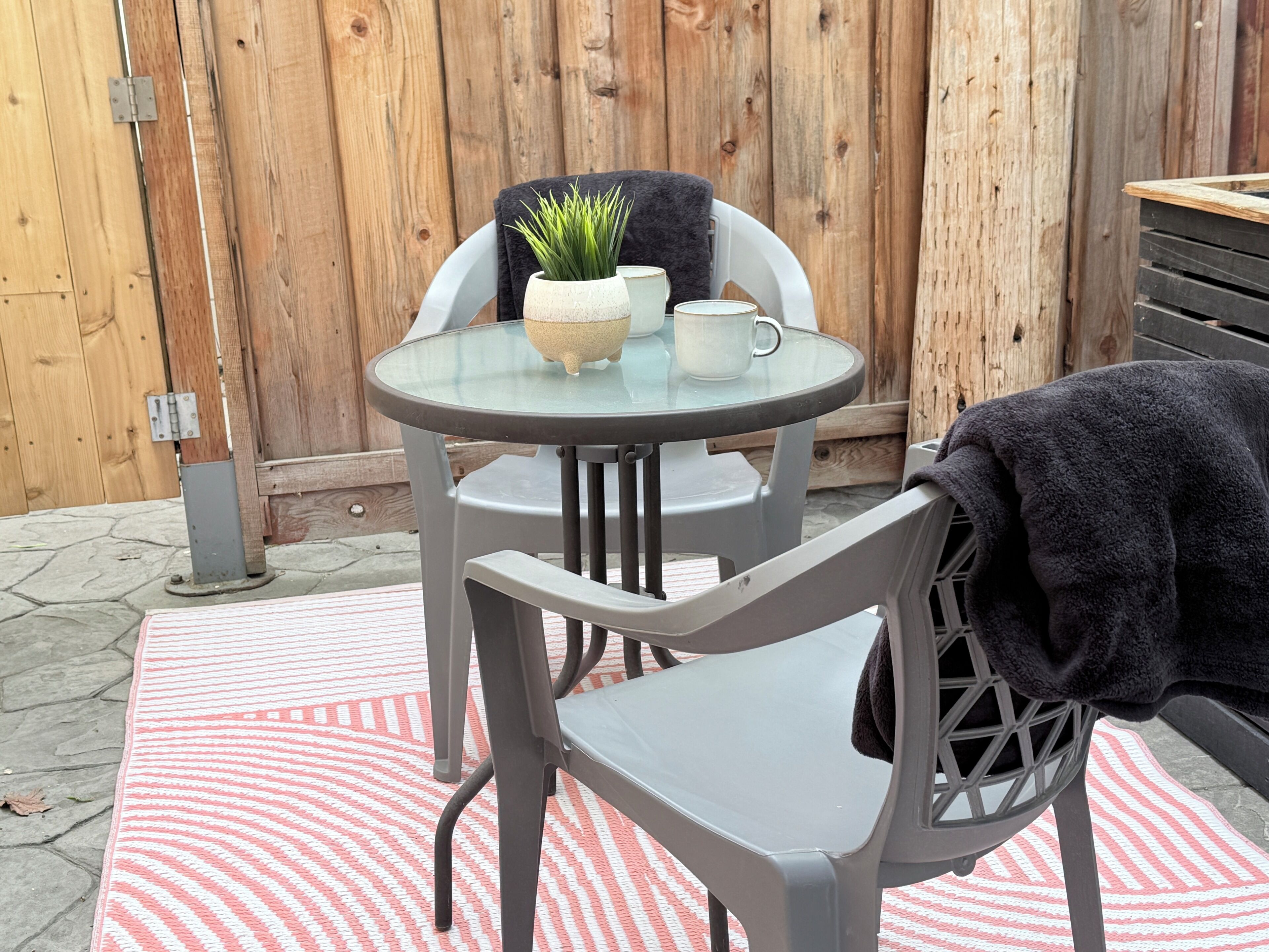 A round glass table with a white bowl and a white mug, two grey chairs, a grey blanket, and a red and white striped rug.