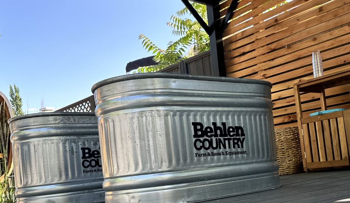 Zwei große Metallbehälter mit der Aufschrift 'Behlen Country Farm & Ranch Equipment', platziert auf einer Holzterrasse mit einem Holzzaun und einem Metalltor im Hintergrund.