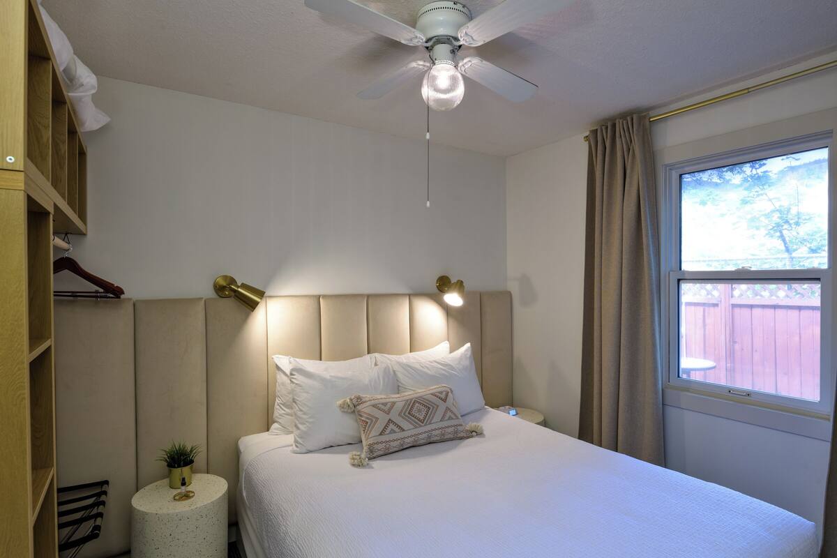 A neatly arranged bedroom with a bed, bedside lamps, a ceiling fan, a window with curtains, and a small round table with a plant.