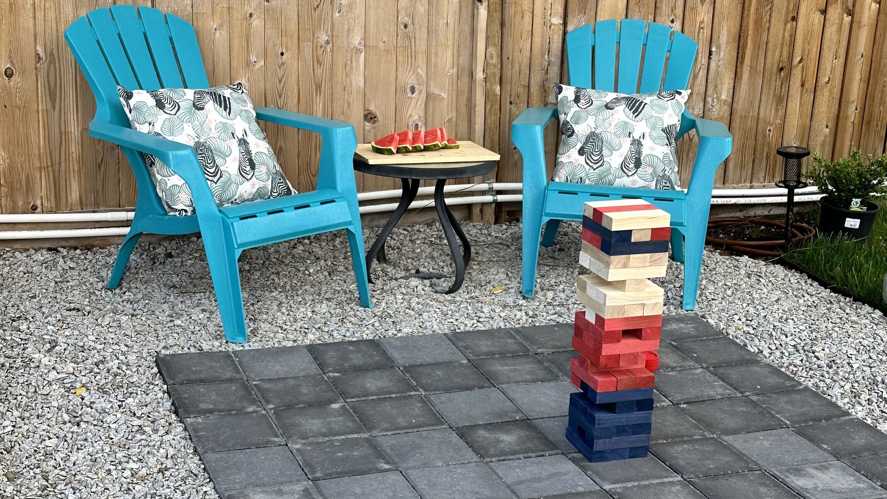 Two blue chairs with patterned cushions, a small round table with a watermelon slice, and a Jenga tower on a stone patio.
