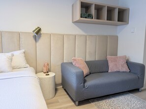 A modern bedroom with a bed, a grey sofa, a small side table, a wall-mounted shelf, and a wooden headboard. - Lakeside Villa & Nordic Spa (Penticton)