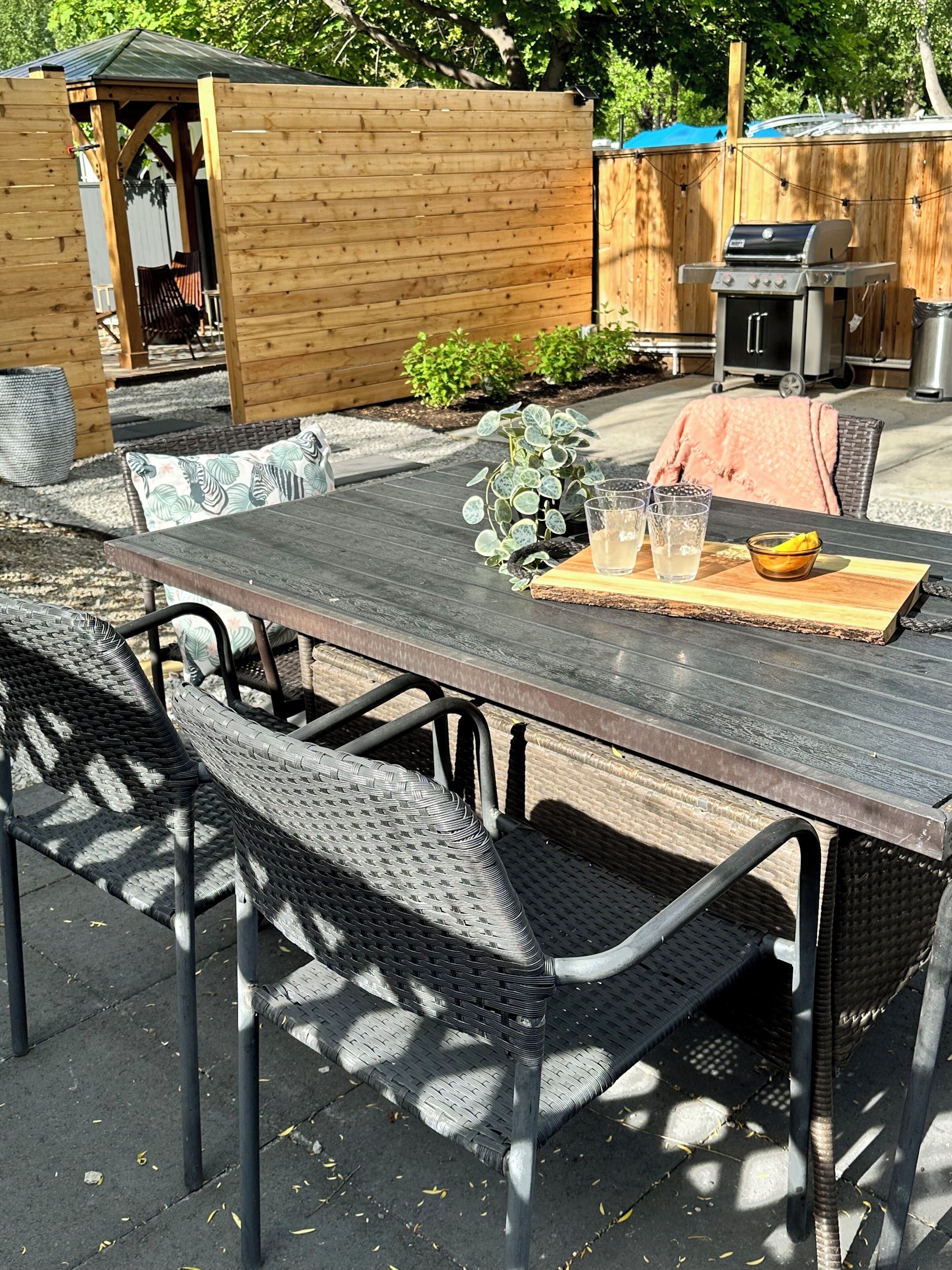 An outdoor patio with a wooden table and wicker chairs, a grill, and a small garden area.
