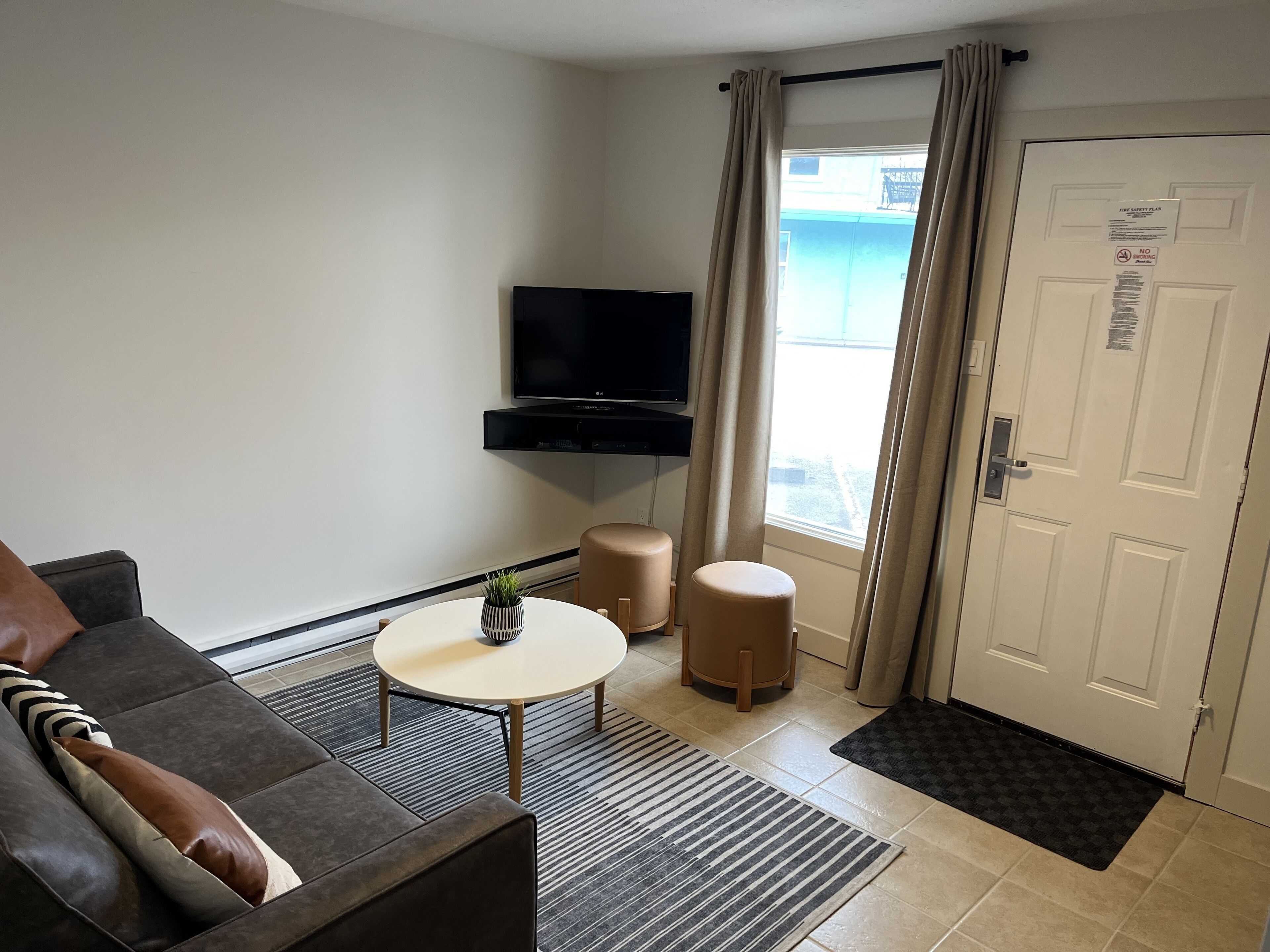 A modern living room with a sofa, a coffee table, a TV mounted on the wall, and a door with a fire safety plan sticker.