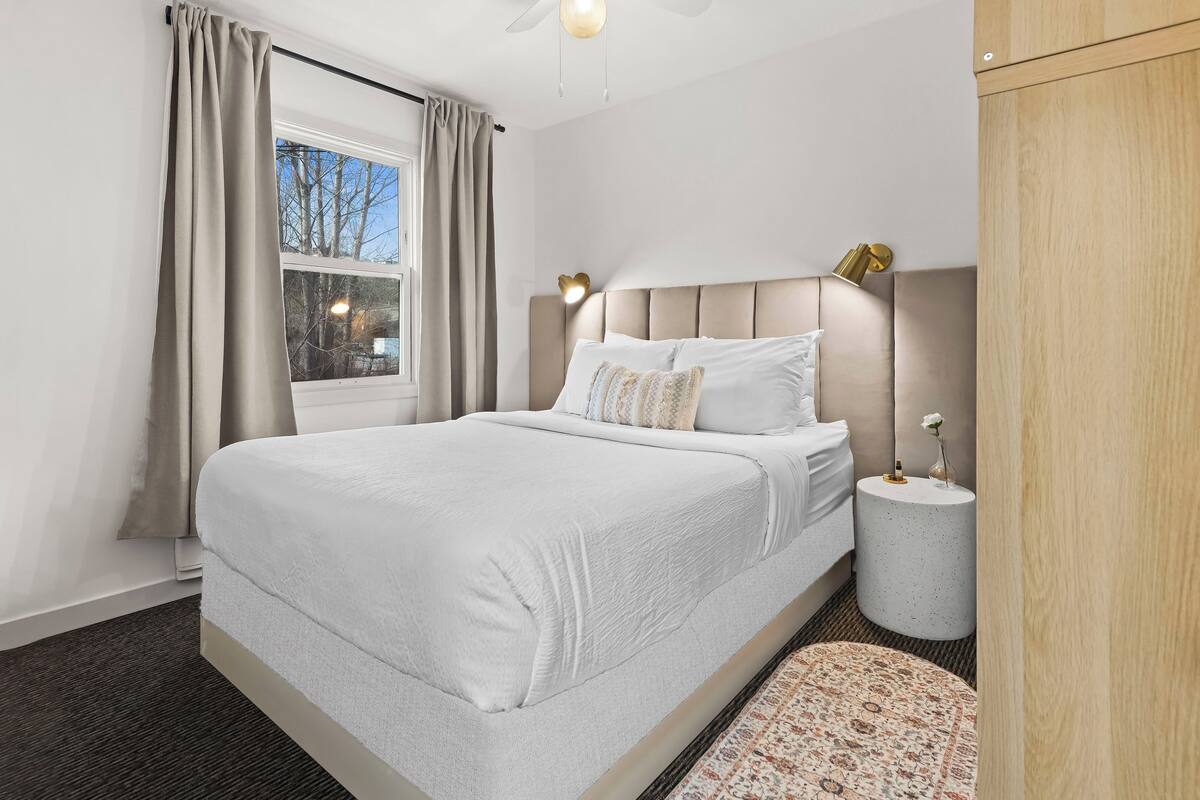 A bedroom with a bed, a window with curtains, a round bedside table with a lamp, and a headboard.