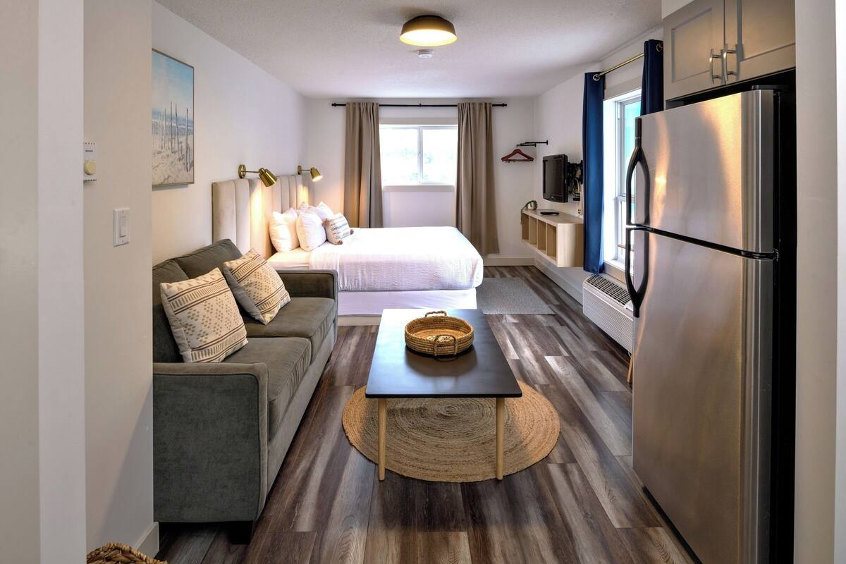 A compact hotel room with a bed, sofa, dining table, and a refrigerator.