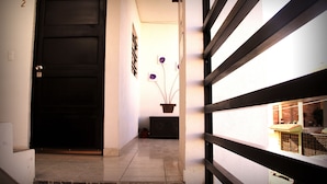 A hallway with a black door, a tiled floor, a potted plant, and a window with a view of a building. - Residencia San Pablo (Querétaro)