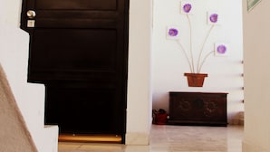 A dark wooden door with a keyhole, a stone-tiled entrance, a small potted plant with purple flowers, and a wooden cabinet. - Residencia San Pablo (Querétaro)