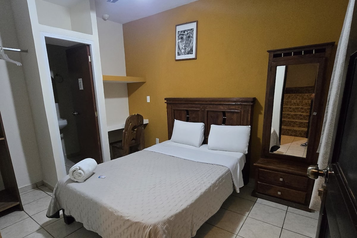 Quarto de hotel com cama, ventilador de teto, espelho e porta para um banheiro.
