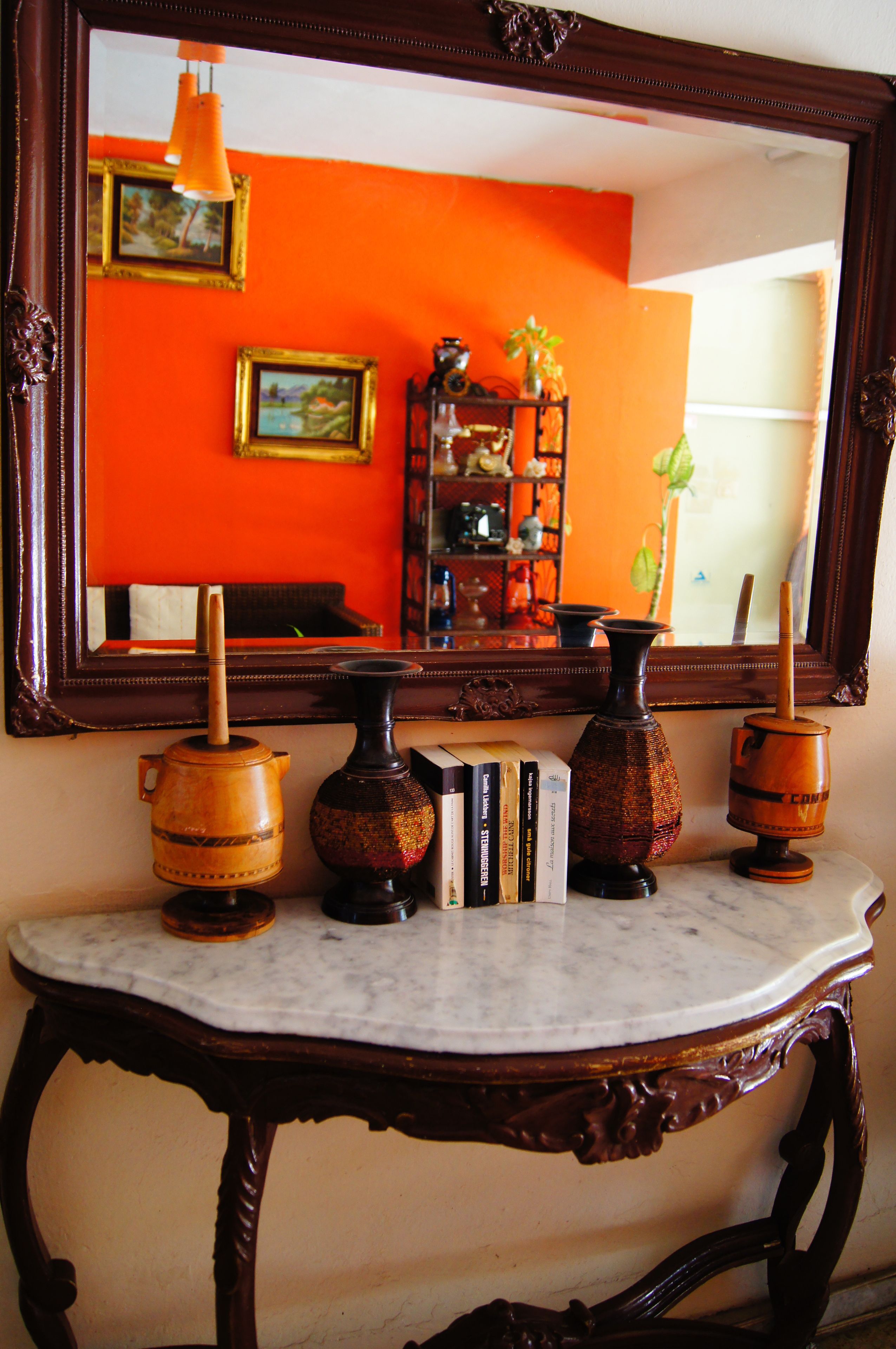 Une console en marbre, agrémentée de poteries décoratives et de livres, est placée devant un mur orange. Un grand miroir reflète une pièce où l’on aperçoit un lit et une plante.