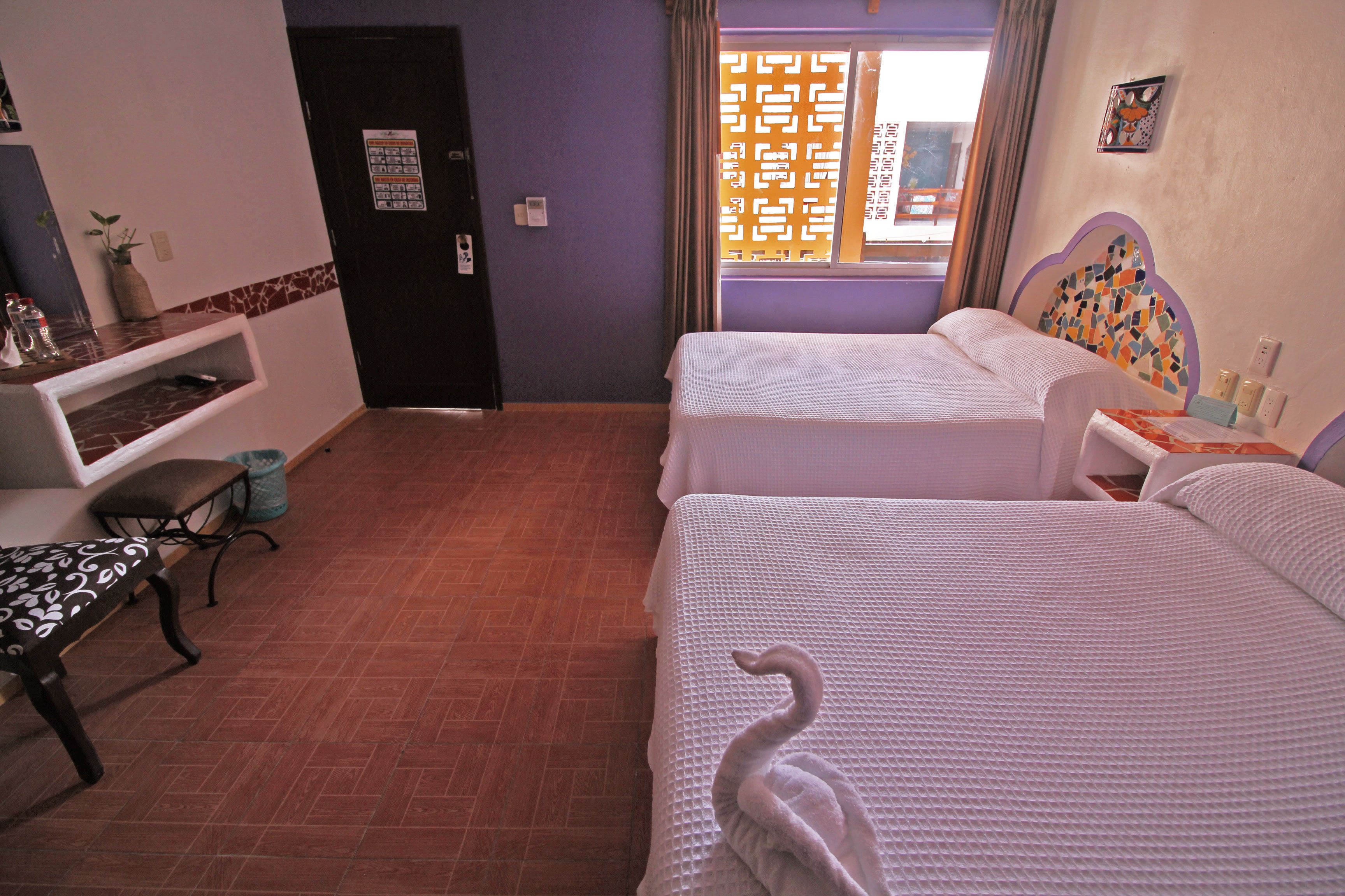 Habitación de hotel con dos camas, un banco, una mesita y una ventana con un panel decorativo.
