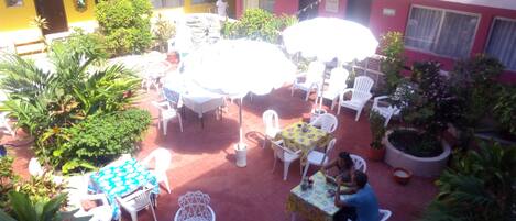 Un patio exterior con sillas y mesas de plástico blanco, rodeado de plantas en macetas y un edificio amarillo al fondo.