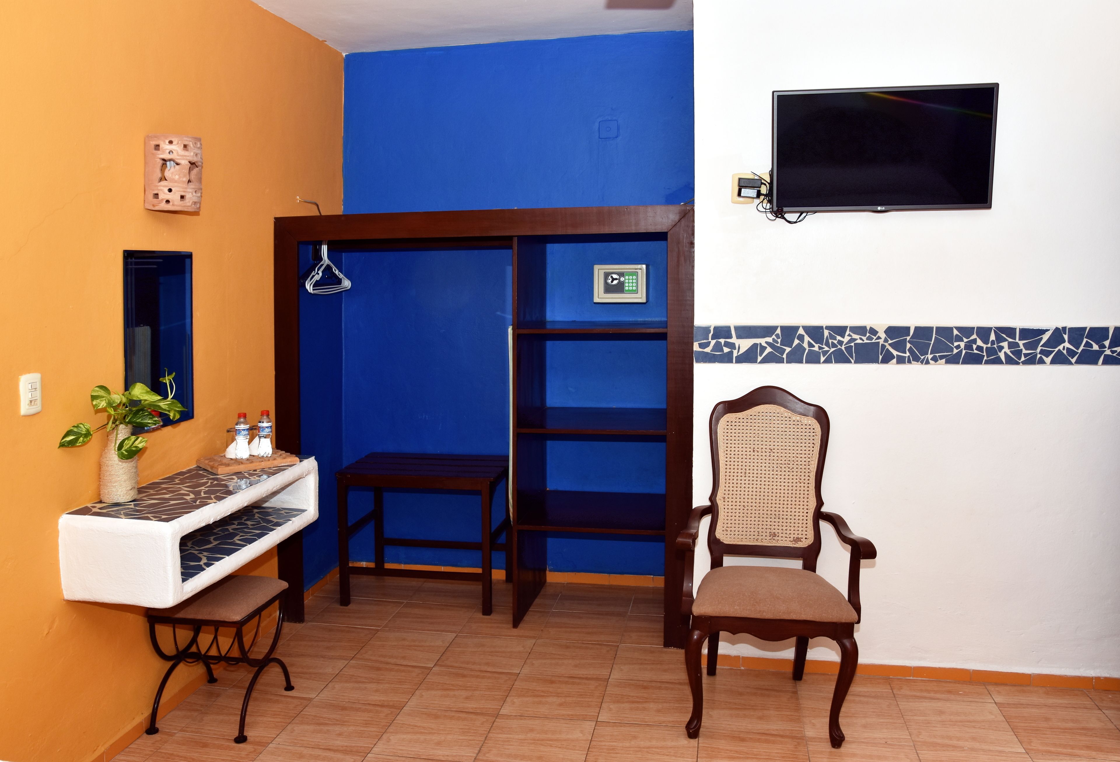 Een kamer met een blauwe wand, een houten stoel, een klein tafeltje, een aan de wand bevestigde televisie en een plank met een fotolijst.