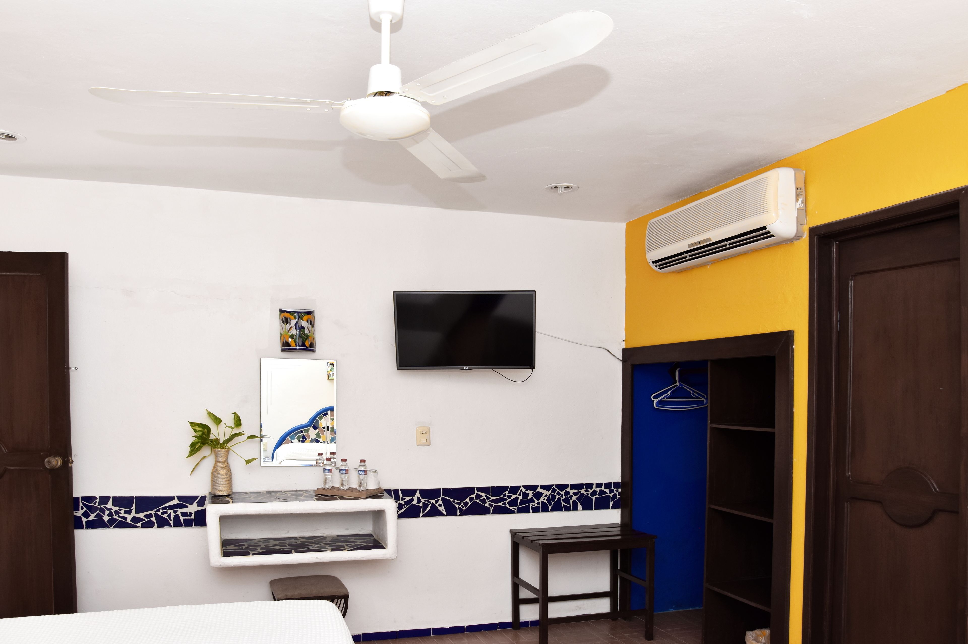 Quarto de hotel com ventoinha no teto, televisão montada, pequena estante com objetos decorativos, planta em vaso e guarda-roupa com interior azul.