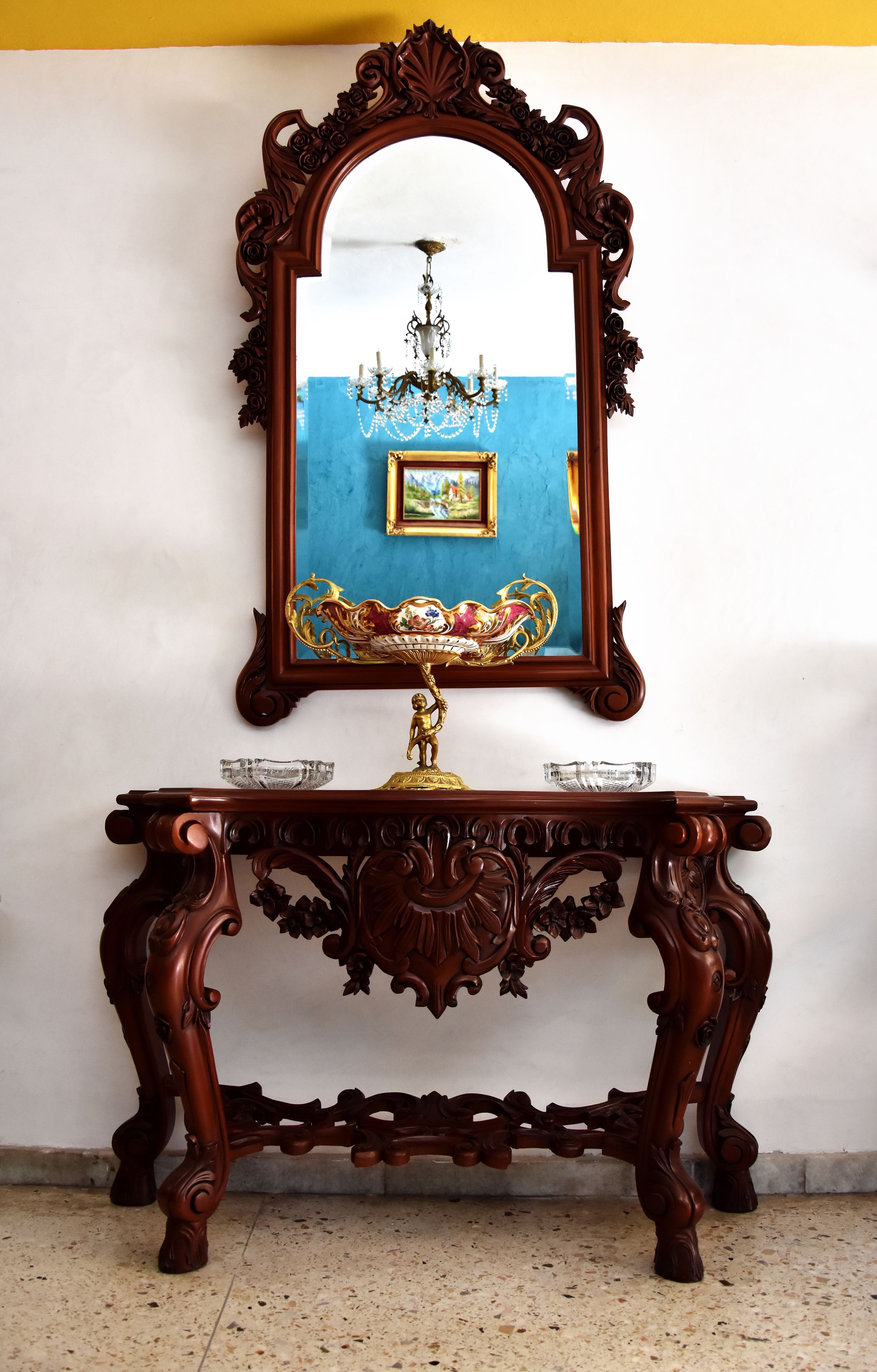 Une console en bois, surmontée d’un miroir qui reflète un lustre et un tableau encadré accroché au mur.