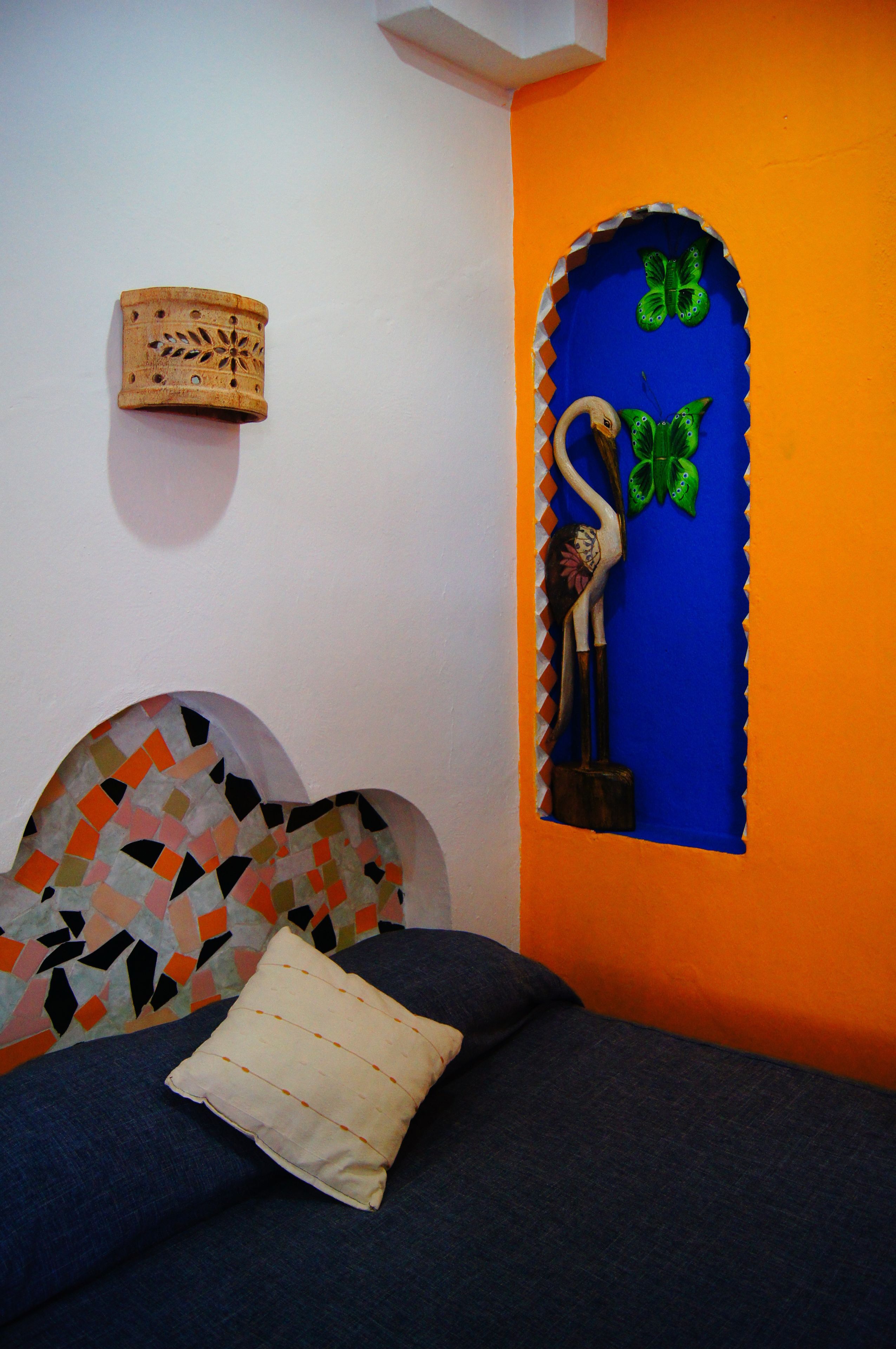 Un dormitorio con una paleta de colores azul y naranja, que incluye una cama con cabecera de estampado geométrico, un adorno de pared y un estante fijado a la pared con una mariposa verde y una escultura de madera.