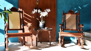 Duas cadeiras de madeira com encostos trançados, uma mesa pequena com um vaso de flores brancas e uma porta de madeira com fechadura.